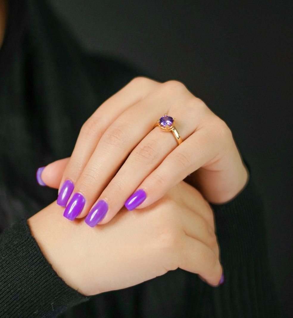 Close-up of woman's hands with vibrant purple nails and a gold ring, set against a dark background.